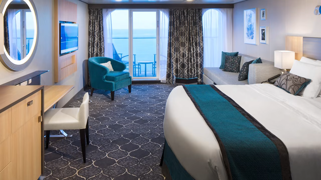 Spacious Royal Caribbean Sea Class suite featuring a king-sized bed with teal accents, a sitting area, and a private balcony overlooking the ocean.